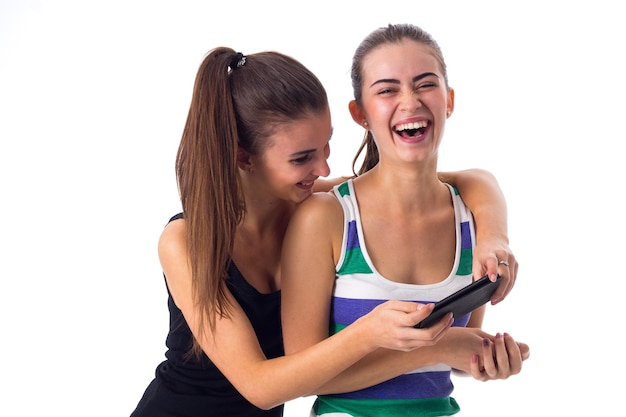 Two smiling young women in striped and black shirts using smartphone and laughing in studio