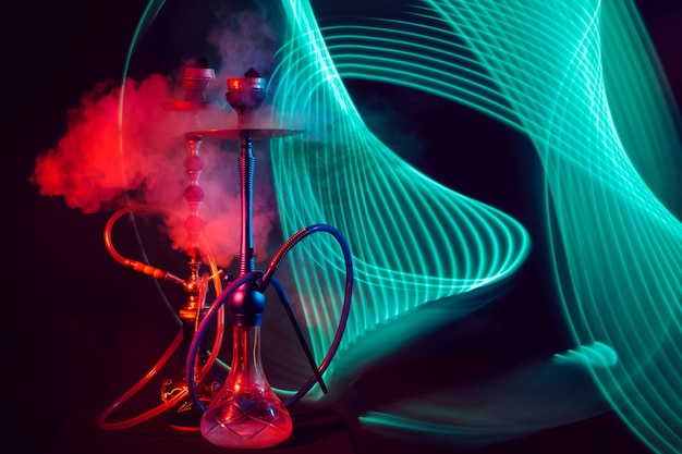 Two smoky hookahs with glass flasks of water with shisha coals with red green neon lighting on a black background