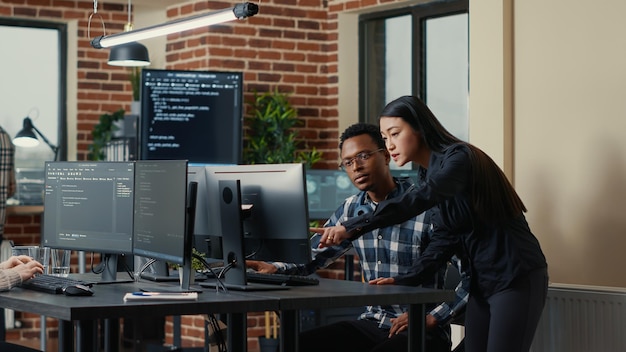 Два разработчика программного обеспечения держат ноутбук с интерфейсом кодирования, идут к столу и садятся, рассказывая о групповом проекте. Команда программистов обсуждает алгоритмы, указывающие на экран компьютера.
