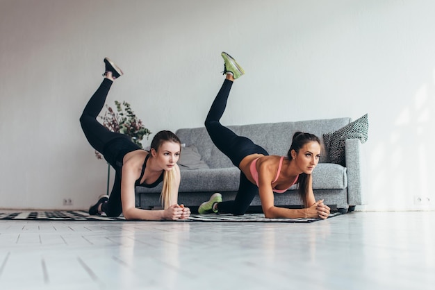 Two sportive female friends doing butt toning exercise performing donkey kicks at home