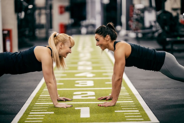 Two sportswomen are doing planks in a gym and smiling at each other