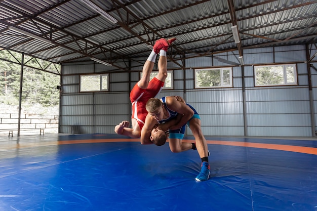 Two strong wrestlers in blue and red wrestling tights are wrestlng  on a wrestling carpet in the gym.