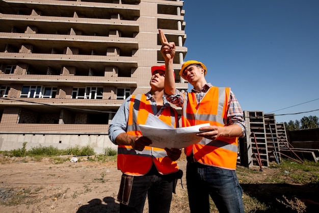 Two structural engineers dressed in shirts, orange work vests and helmets explore construction documentation against the background of a multistorey building .