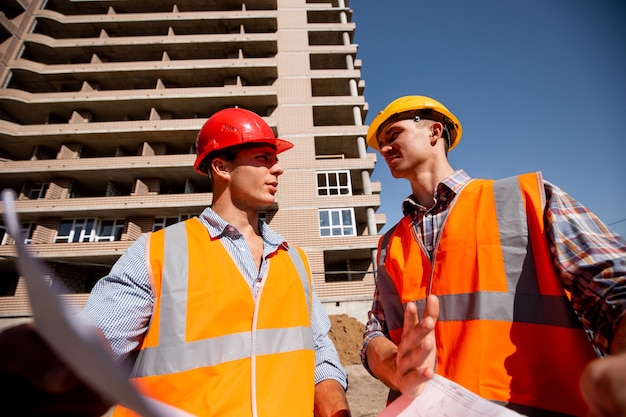 Two structural engineers dressed in shirts, orange work vests and helmets explore construction documentation against the background of a multistorey building .