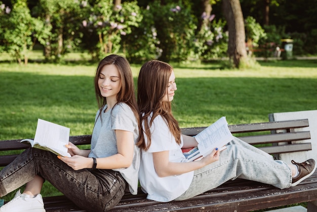 Две девушки-студентки смотрят на раскрытую книгу на скамейке в парке. Дистанционное обучение, подготовка к экзаменам. Мягкий выборочный фокус.