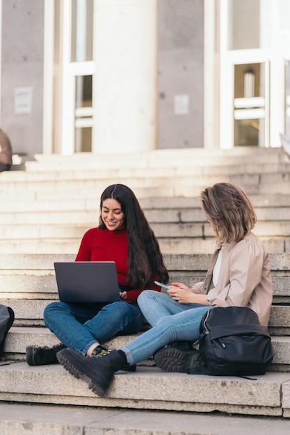 Two student girls sitting on the stairs of the college with a laptop and mobile