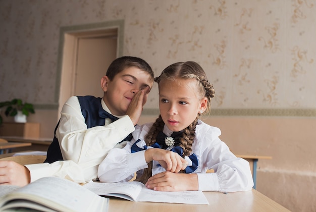 Foto due studenti, un ragazzo e una ragazza, siedono a una scrivania a scuola e comunicano tra loro nell'orecchio.