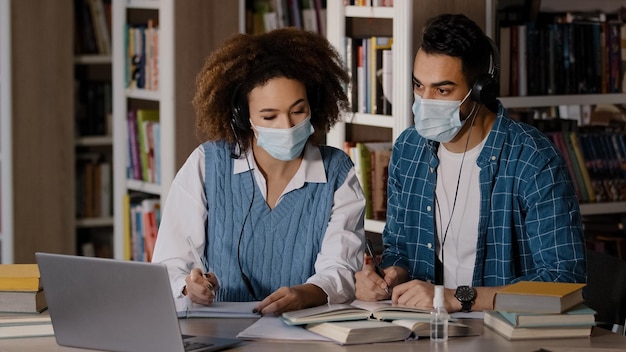 의료 마스크를 입은 두 명의 학생이 대학 도서관의 책상에 앉아 헤드폰을 착용한 교사를 듣고 있습니다.