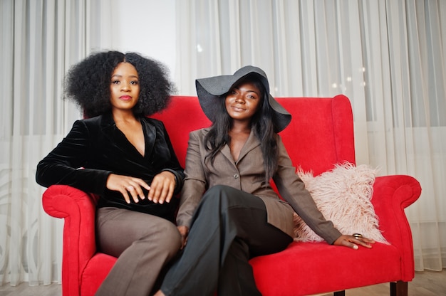 Two stylish african american womans in formal wear and hat sitting on red couch at white room.