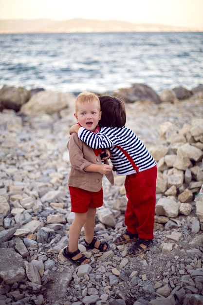 Two stylish baby boys in vests stand on the seashore of pebbles in summer