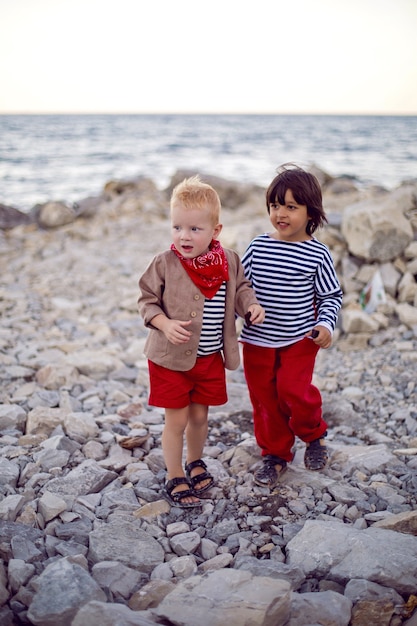 Two stylish baby boys in vests stand on the seashore of pebbles in summer