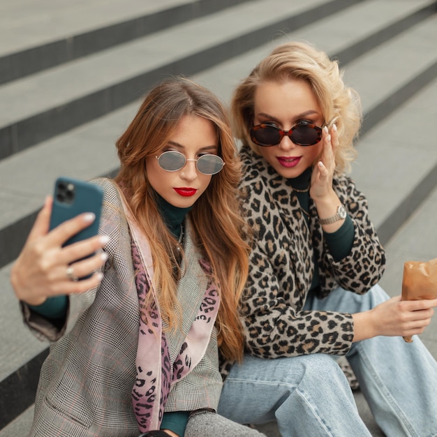 Two stylish bright girls in fashionable clothes sit on the steps and take a selfie photo on the phone