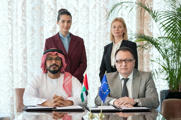 Foto due partner commerciali di successo o delegati in abiti da cerimonia seduti al tavolo davanti alla telecamera contro eleganti donne d'affari nella sala riunioni