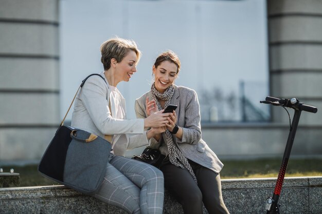 A two successful businesswomen surfing on smartphone while having a quick break in front of the office building.