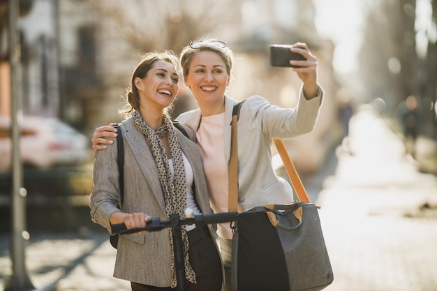 두 명의 성공적인 경제인이 도시를 걸으면서 스마트폰으로 셀카를 찍고 있습니다.