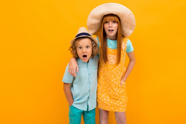 Two surprised red-haired brother and sister are looking in summer hats,  on a yellow