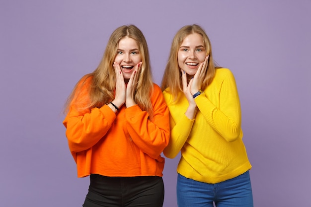 Two surprised young blonde twins sisters girls in vivid colorful clothes putting hands on cheeks isolated on pastel violet blue wall  . People family lifestyle concept. .