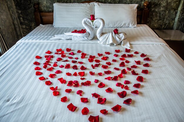 Photo two swans on the bed rolled from towels on a white bed with red roses arranged in a heart shape special decorations for honeymoon and valentines day in hotel