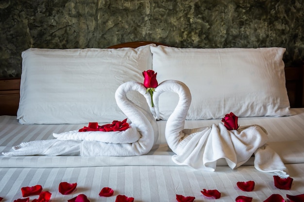 Two swans on a bed rolled from towels on a white bed with red roses specially decorated for honeymoon and valentines day in hotel