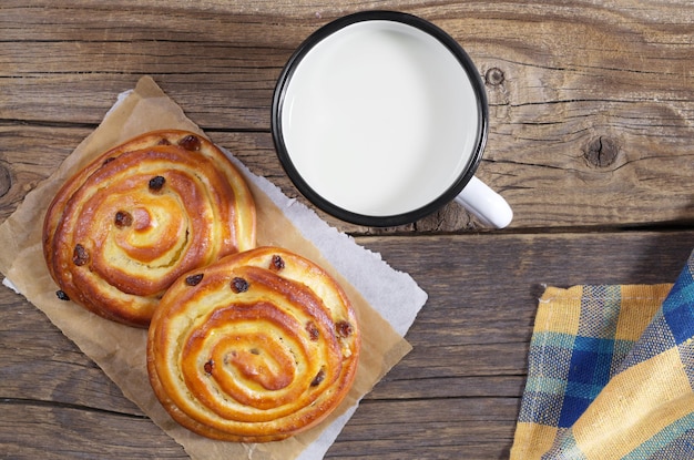 Two sweet buns with raisins and enamel mug of milk on rustic wooden table, top view