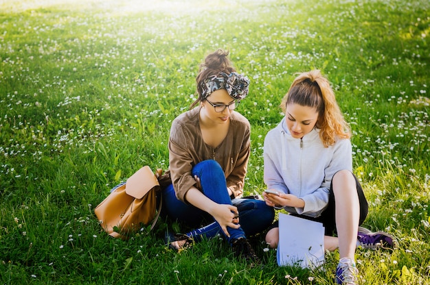 Two sylish girls sitting on the grass and using phone