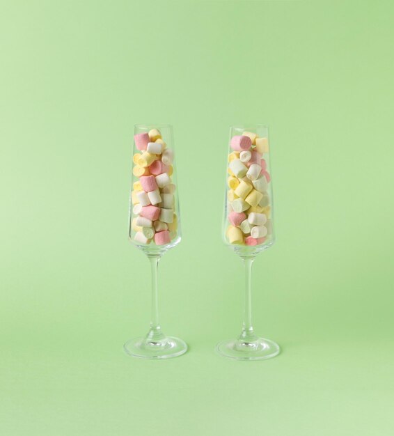 Two tall glass glasses filled with colorful marshmallows on a green background. The concept of happiness and success.