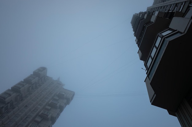 Two tall residential buildings in the form of a tower, immersed in a misty gray sky. Cyberpunk stylistics. Dramatic, depressing mood.