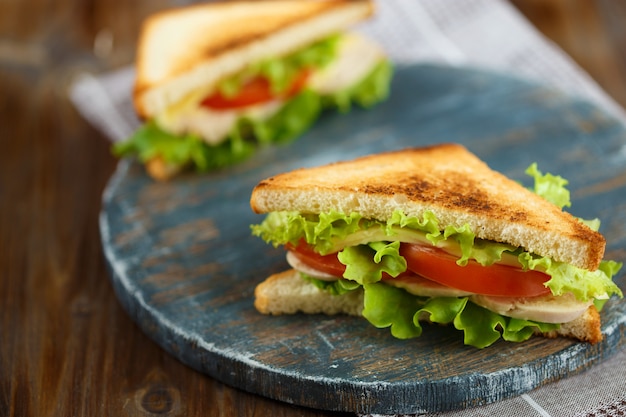 Two tasty sandwich with chicken, tomatoes, lettuce, cheese on a wooden plate on a dark background