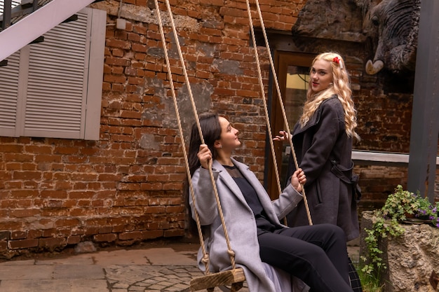 Two teen girls are talking in the courtyard of an old building, one of them is sitting on a swing