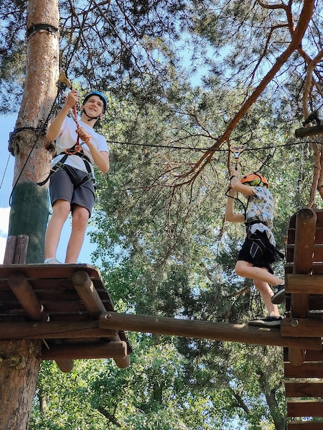 Two teenage boys are standing on a wooden pallet on a tree in a rope park in a safety harness