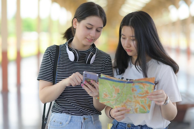 Two teenage women holding smart phones and maps to travel,\
foreign teenage girls holding smart phones, asian teenage girls\
holding maps, travel planning, front photo.