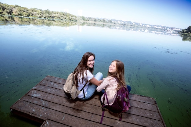 写真 川岸の桟橋と背景の街に座るバックパックを持つ 2 人の 10 代の女の子