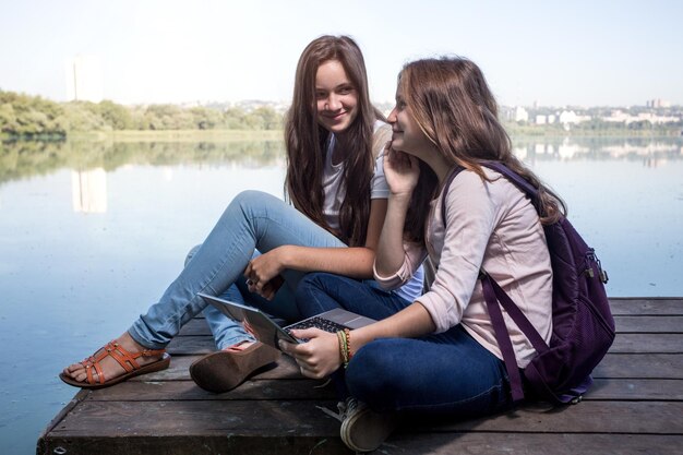 Photo two teenager girls with backpacks sitting on a pier at the river bank and the city in the background