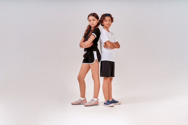 Two teenagers cute boy and girl in sportswear looking at camera while posing isolated over grey