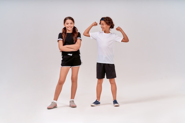 Two teenagers having workout happy boy showing his biceps to a cute smiling girl standing isolated