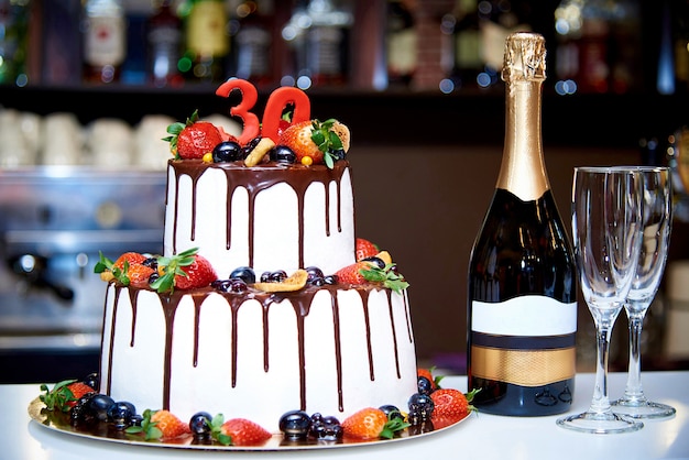 A two-tiered white cake with fresh fruit and chocolate stands next to a bottle of champagne