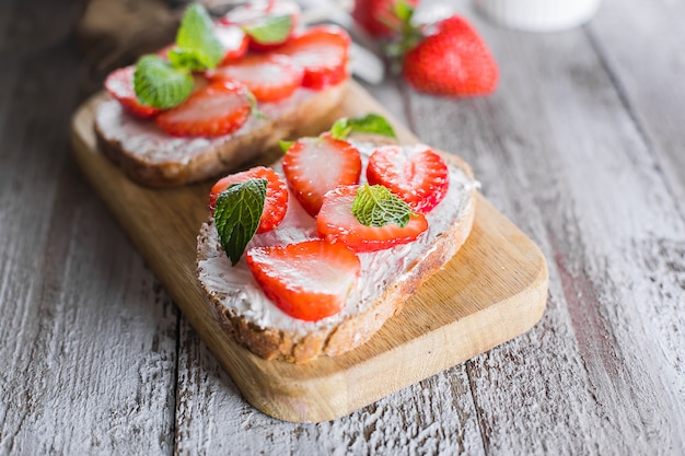 Two toasts or bruschetta with strawberry and mint on cream-cheese on wooden board on table