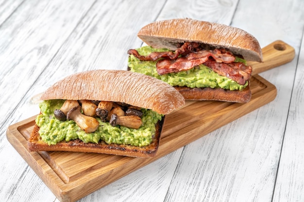 Two toasts with guacamole, mushrooms and bacon on cutting board