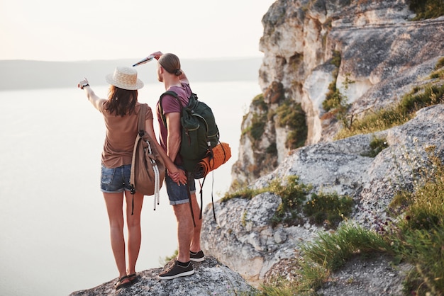 Two tourist male and woman with backpacks stand to the top of the crag and enjoying sunrise. Traveling mountains and coast, freedom and active lifestyle concept