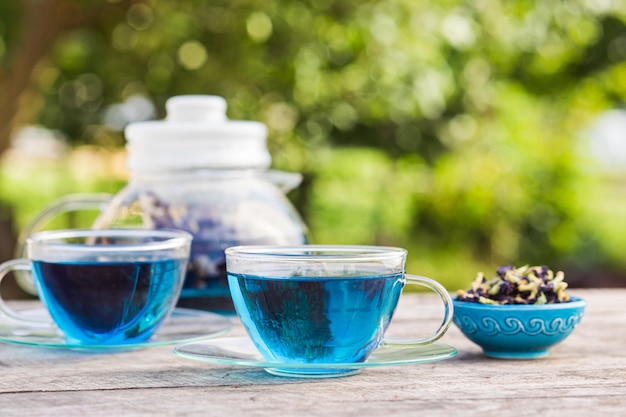 Two transparent cups of blue tea a small plate with dried\
butterfly pea flower and a teapot in the background stand on a\
wooden table