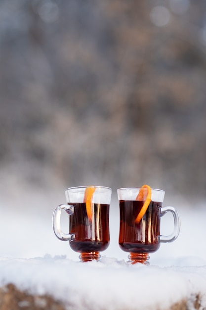 Two transparent glasses with wine and orange slices in the forest on a snowy tree