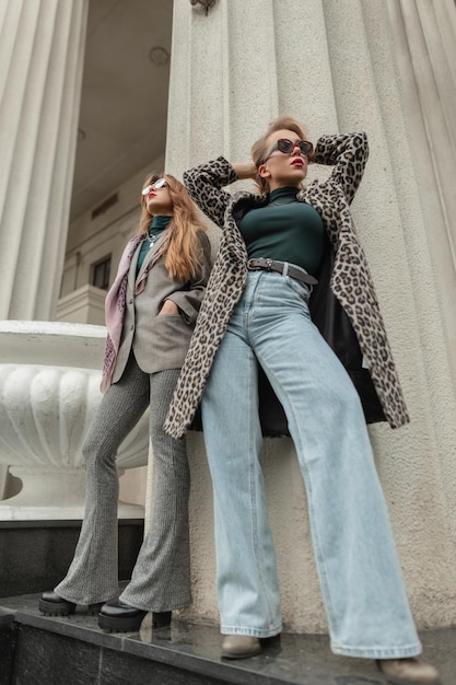 Due giovani donne alla moda in abiti eleganti autunnali con cappotto leopardato, scarpe jeans blu campana e occhiali da sole, si ergono e posano vicino a colonne vintage
