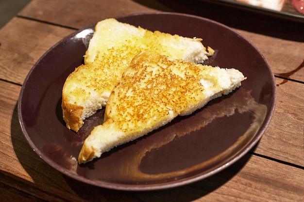 Two triangular slices of baked garlic butter bread are placed on a dark brown porcelain plate on a wooden table