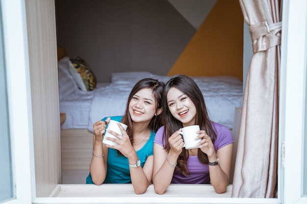 Two twin Attractive asian sister holding a cup with hot tea or coffee near the window in her home