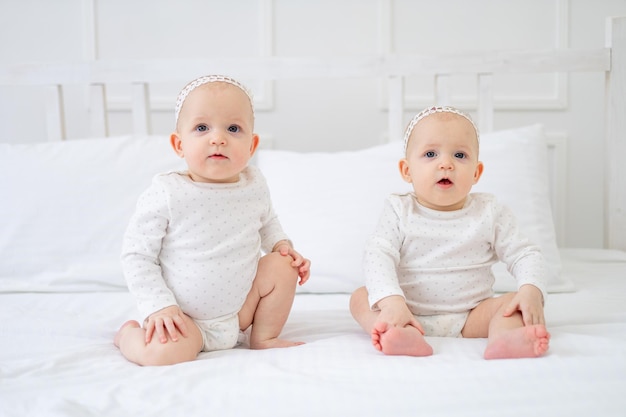 Two twin babies girls of six months on a white cotton bed in a bodysuit on the bed at home playing and smiling