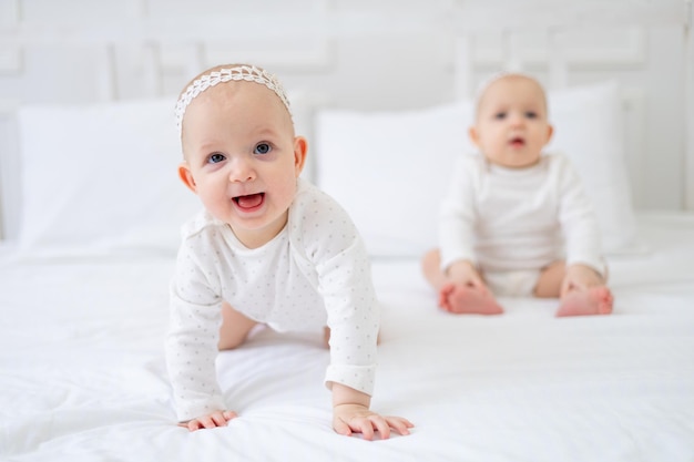 Two twin babies girls of six months on a white cotton bed in a bodysuit on the bed at home playing and smiling