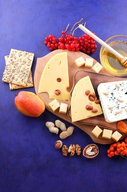 Two types of cheese with fruits berries nuts and dried fruits on a wooden board Various types of cheese composition with honey and bread rolls on a blue background