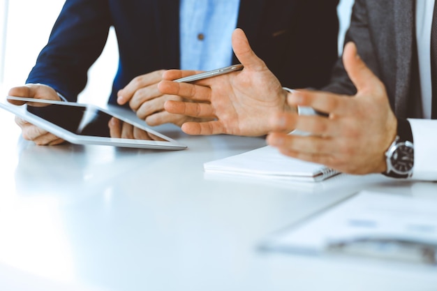 Two unknown businessmen or partners using tablet pc and discussing questions at meeting in modern office, close-up of hands. Group of managers at negotiation or brainstorm. Teamwork, partnership and b