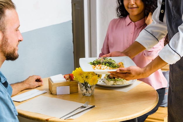 Two unrecognizable people eating in a restaurant and the server placing the food on the table.