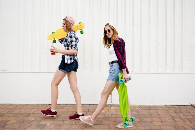 Two urban girls with longboards wearing checkered shirts going along the street in the city.